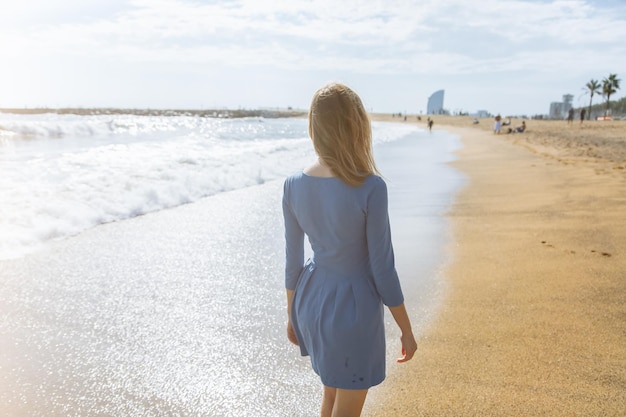 Schönes Mädchen im blauen Kleid geht am Strand Erstaunliches Sommerfoto Frau in der Nähe des Meeres Fröhliche und lustige Emotionen Urlaubsreisekonzept Schlanke Beine Warmes Meerwasser