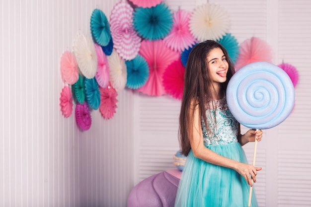 Schönes Mädchen im blauen Kleid hält große Süßigkeiten in ihren Händen und zeigt Zunge lächelnd. Porträt des Mädchens im Studio mit Dekoration der Makrone.