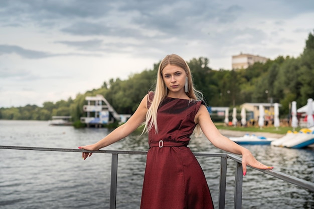 Schönes Mädchen im langen, modischen, dunkelroten Kleid in der Nähe des Teichs im Stadtpark-Lifestyle