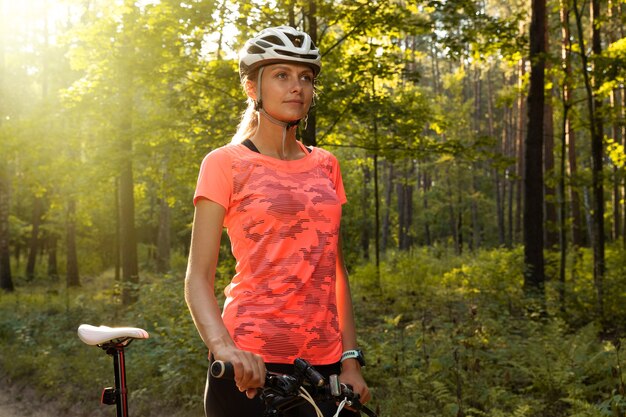 Schönes Mädchen im leuchtend orangefarbenen T-Shirt, in den Shorts und im Helm reitet im Wald auf einem Fahrrad