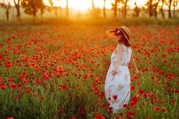 Schönes Mädchen im Mohnfeld bei Sonnenuntergang in einem weißen Kleid und Hut