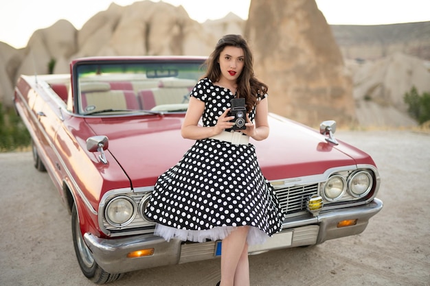 Schönes Mädchen im Retro-Stil, das in der Nähe eines roten Cabriolet-Oldtimers mit alter Filmkamera in den Händen posiert
