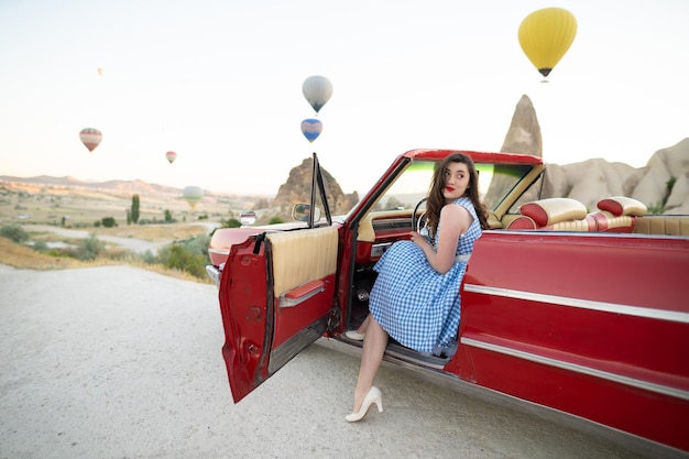 Schönes Mädchen im Retro-Stil, das neben einem roten Cabriolet-Oldtimer auf dem Hintergrund von Luftballons in Kappadokien posiert