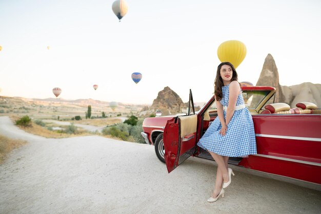 Schönes Mädchen im Retro-Stil, das neben einem roten Cabriolet-Oldtimer auf dem Hintergrund von Luftballons in Kappadokien posiert