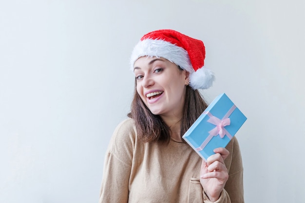 Schönes Mädchen im roten Weihnachtsmann-Hut, der in der Hand blaue Geschenkbox lokalisiert auf weißem Hintergrund hält. Porträt der jungen Frau, wahre Emotionen. Frohes Weihnachts- und Neujahrskonzept.