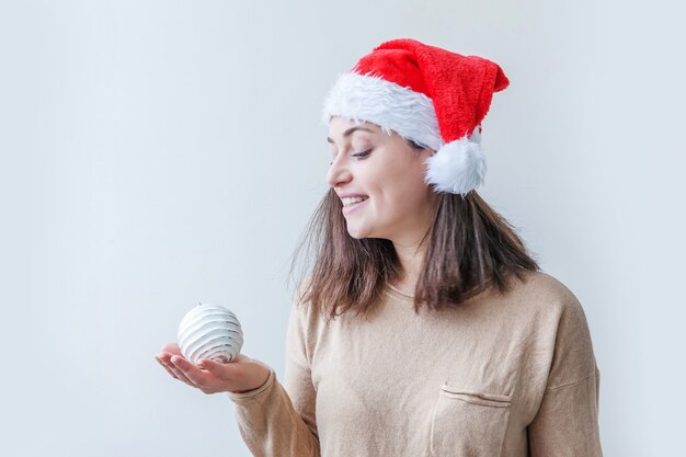 Schönes Mädchen im roten Weihnachtsmann-Hut, der Weihnachtsbaumverzierungsball in der Hand lokalisiert auf weißem Hintergrund hält. Porträt der jungen Frau, wahre Emotionen. Frohes Weihnachts- und Neujahrskonzept.