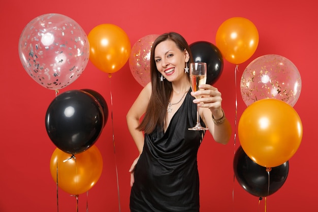 Schönes Mädchen im schwarzen Kleid feiernd, Hand mit Glas Champagner auf Kamera auf leuchtend roten Hintergrundluftballons zeigend. Frauentag, Frohes neues Jahr, Geburtstagsmodell-Feiertagsparty-Konzept.
