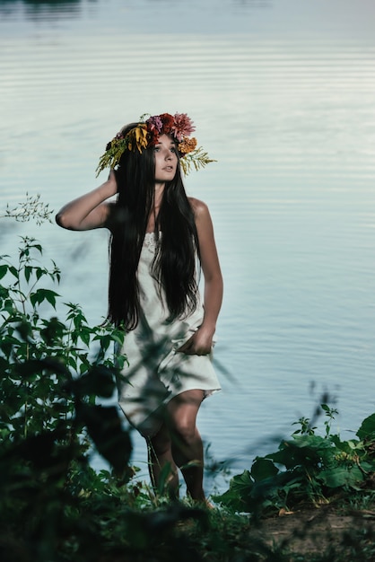 Schönes Mädchen im weißen Weinlesekleid und im Kranz von Blumen am Wasser