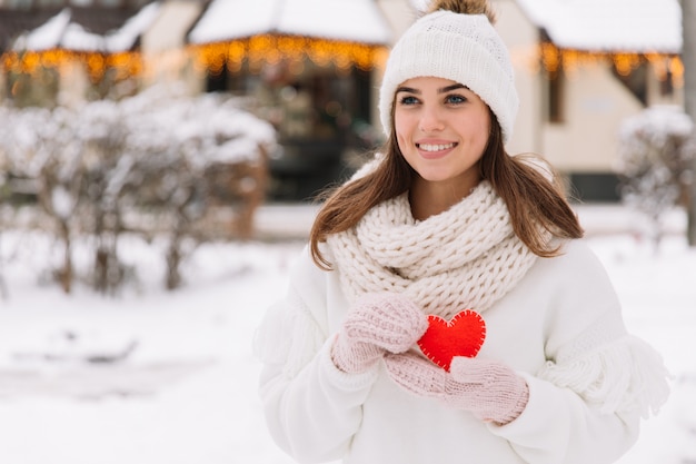 schönes Mädchen in der weißen Winterkleidung auf der Straße, die Herz zum Valentinstag hält