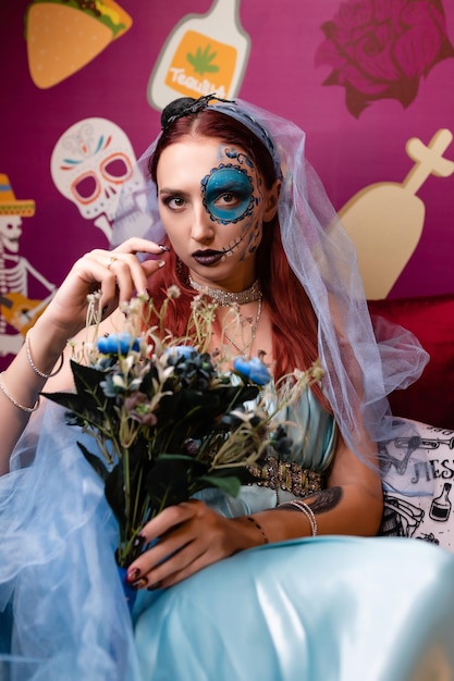 Schönes Mädchen in einem blauen Kleid mit künstlerischem Make-up auf ihrem Gesicht hält einen Blumenstrauß Posing beim Sitzen Halloween-Maske Halloween-Konzept Nachtclub-Maskerade
