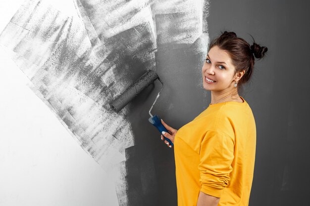 Schönes Mädchen in einem gelben Pullover malt eine Wand in grauer Farbe. Raummalerei, Reparatur, Design. Speicherplatz kopieren.