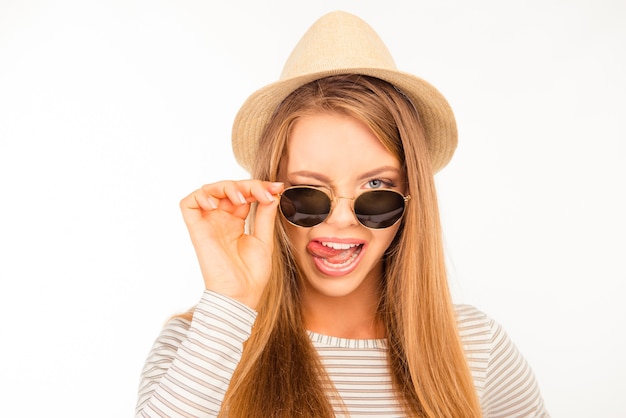 Schönes Mädchen in einem Hut und einer Sonnenbrille, die ihre Zunge zeigt