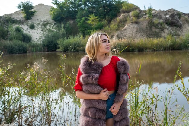 Schönes Mädchen in einem Pelzmantel und einem hellroten T-Shirt und Shorts, die am See an einem warmen Herbsttag aufwerfen