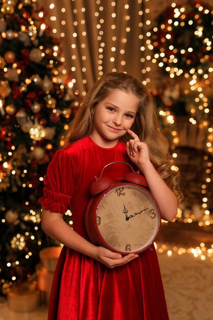 Schönes Mädchen in einem roten Kleid in einer Weihnachtsdekoration mit einer großen Uhr in ihren Händen