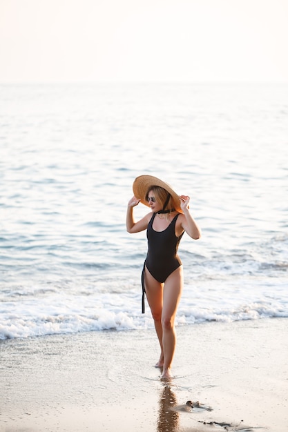 Schönes Mädchen in einem schwarzen Badeanzug und Hut an einem Sandstrand am Meer im Sonnenuntergangssonnenlicht