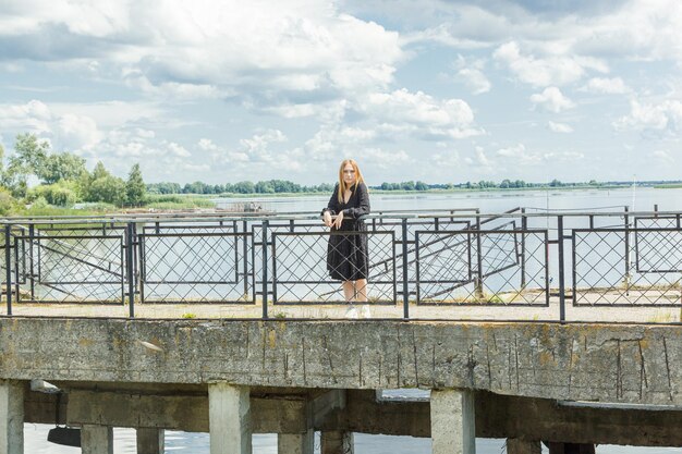 Schönes Mädchen in einem schwarzen Kleid auf dem Hintergrund des Flussmeeres mit blauem Himmel und Wolken