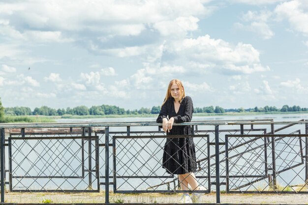 Schönes Mädchen in einem schwarzen Kleid auf dem Hintergrund des Flussmeeres mit blauem Himmel und Wolken