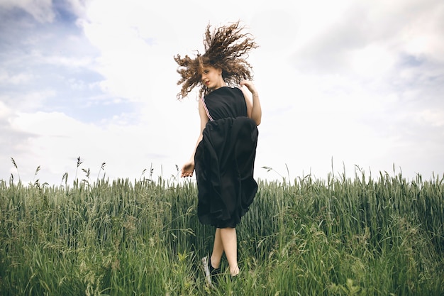 Schönes Mädchen in einem schwarzen Kleid, das im grünen Feld steht und ihr Haar flattert