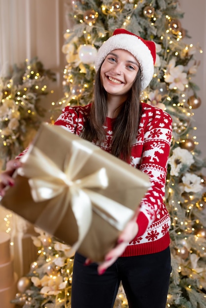 Schönes Mädchen in einem Weihnachtspullover und einem roten Weihnachtsmann-Hut sitzt in der Nähe eines luxuriösen Weihnachtsbaums Lächelnd Hält ein Geschenk in seinen Händen Feier des neuen Jahres, Weihnachten