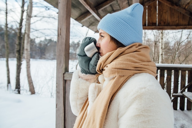 Schönes Mädchen in einem weißen Pelzmantel trinkt warmen Tee im Winter in einem verschneiten Wald