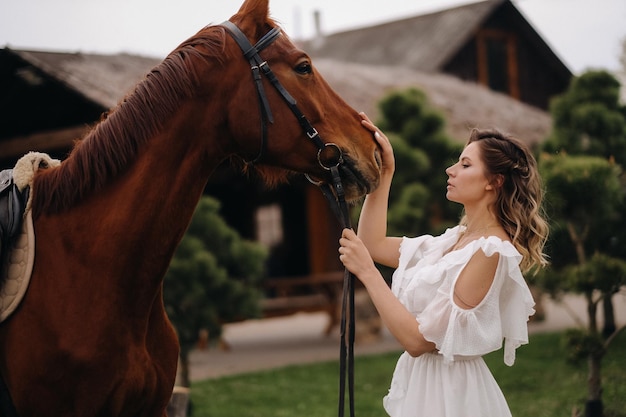 Schönes Mädchen in einem weißen Sommerkleid neben einem Pferd auf einer alten Ranch