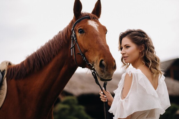 Schönes Mädchen in einem weißen Sommerkleid neben einem Pferd auf einer alten Ranch