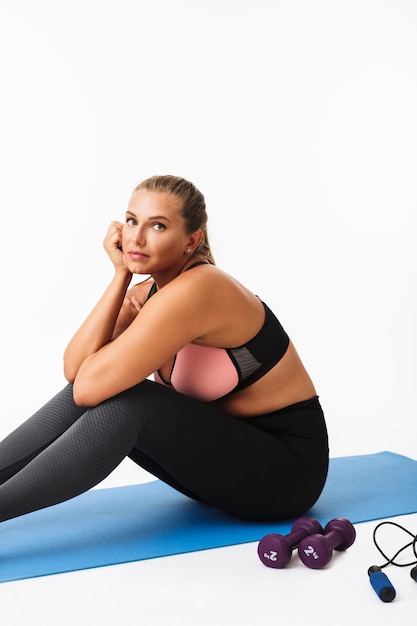 Schönes Mädchen mit Übergewicht in sportlichem Top und Leggings auf Yogamatte sitzend, während sie verträumt vor weißem Hintergrund in die Kamera schaut