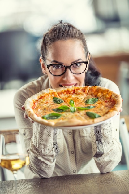 Schönes Mädchen mit Brille beißt in eine runde ganze Pizza in ihren Händen