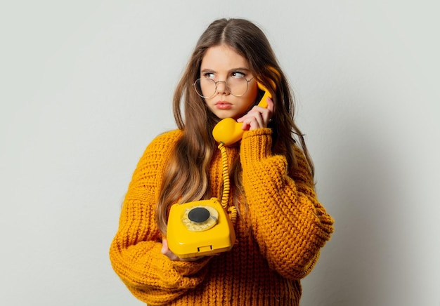 Schönes Mädchen mit Brille und gelbem Pullover mit gelbem Telefon auf weißem Hintergrund