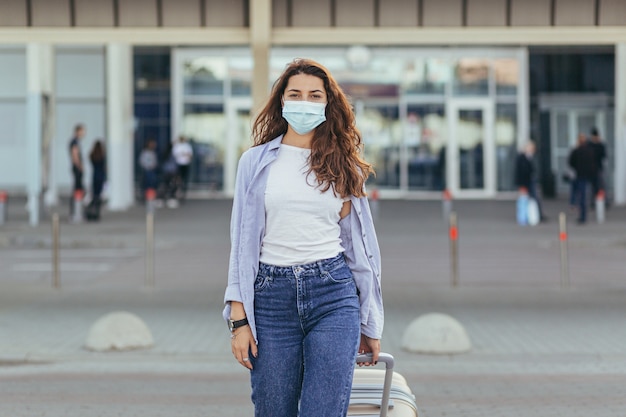 schönes Mädchen mit einem großen Koffer am Flughafen, der eine Schutzmaske auf ihrem Gesicht trägt