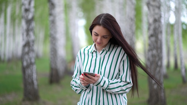Schönes Mädchen mit einem Smartphone in ihren Händen geht entlang eines Birkenhains im Frühjahr.