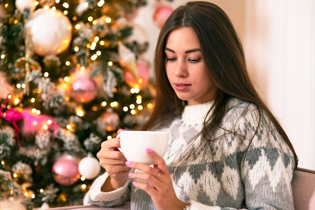 Schönes Mädchen mit einer Tasse in den Händen begrüßt das neue Jahr und Weihnachten am Baum. Mädchen zu Hause, einen Drink gucken, gemütlich