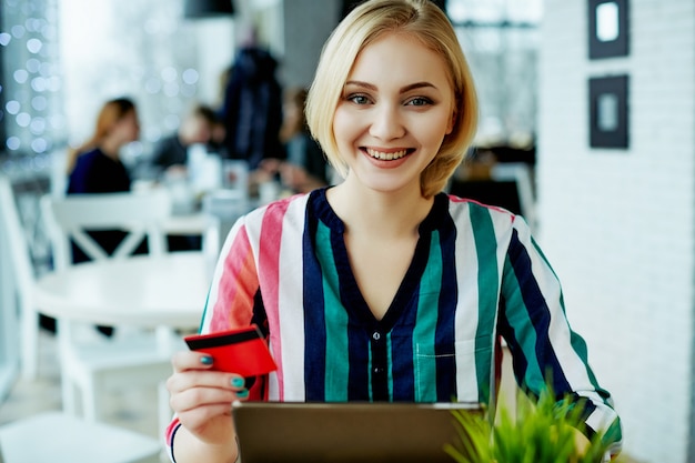 Schönes Mädchen mit hellem Haar, das buntes Hemd trägt, das im Café mit Tablette, Kreditkarte und Tasse Kaffee, freiberufliches Konzept, Online-Einkauf sitzt.
