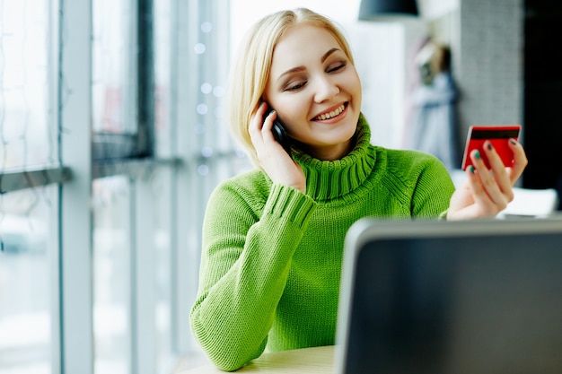 Foto schönes mädchen mit hellem haar, das grünen pullover trägt, sitzt im café mit laptop und kreditkarte, hält handy, porträt, freiberufliches konzept, online-einkauf.