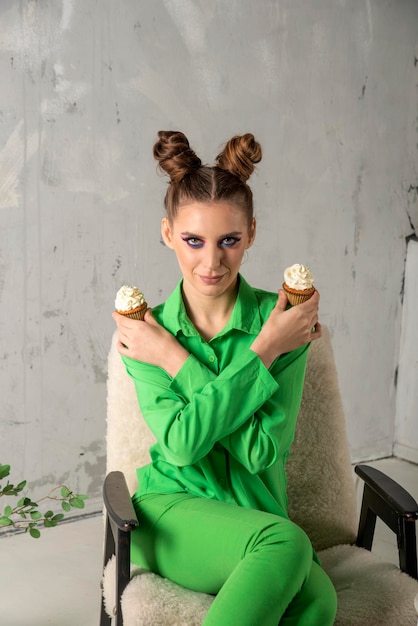 Schönes Mädchen mit hellem Lidschatten und Brötchenfrisur hält zwei Cupcakes mit Sahne Junge Frau in Grün auf grauem Hintergrund