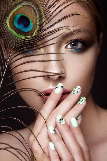 Schönes Mädchen mit hellem Make-up-Maniküre-Design und Pfauenfeder auf ihrem Gesicht Kunstnägel