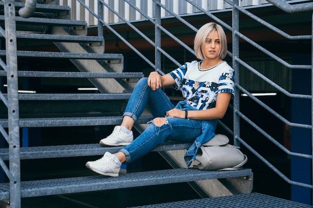 Schönes Mädchen mit kurzen weißen Haaren in Jeans im urbanen Stil sitzt auf der Metalltreppe