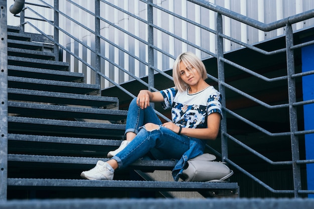 Schönes Mädchen mit kurzen weißen Haaren in Jeans im urbanen Stil sitzt auf der Metalltreppe