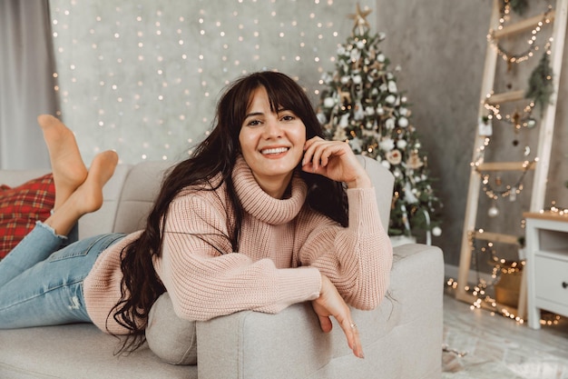 Schönes Mädchen mit langen dunklen Haaren liegt auf dem Sofa vor dem Hintergrund des Weihnachtsbaums. Weihnachten