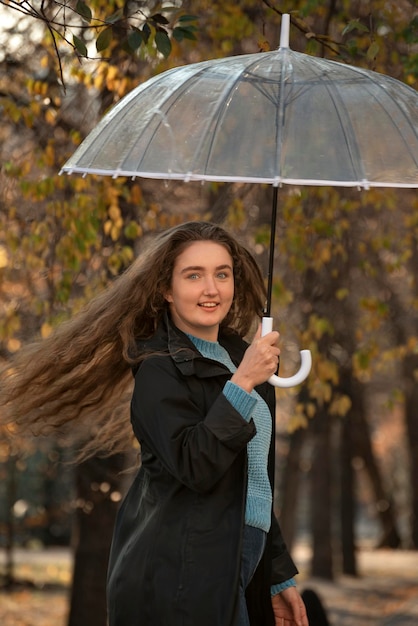 Schönes Mädchen mit langen Haaren, das im Herbstpark unter einem transparenten Regenschirm wirbelt Frau lächelt direkt in die Kamera