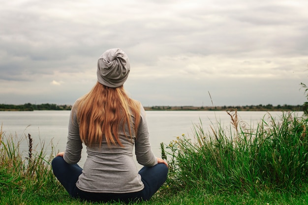 Schönes Mädchen mit langen Haaren sitzt am Ufer. Der Blick von hinten. Sonnenuntergang. Frieden und Ruhe. Yoga am See