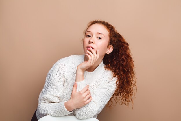 Schönes Mädchen mit langen roten Locken in einem weißen Pullover im Studio auf beigem Hintergrund