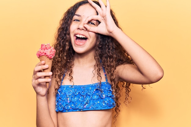 Schönes Mädchen mit lockigem Haar, das einen Bikini trägt, der Eis hält, lächelt glücklich und macht ein Ok-Zeichen mit der Hand auf dem Auge, das durch die Finger schaut