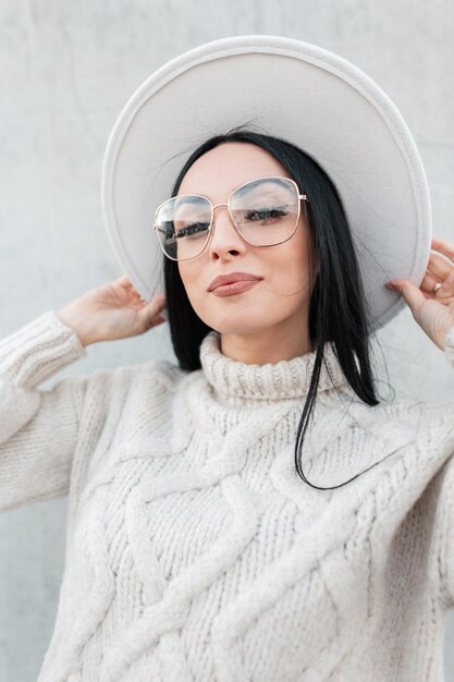 Schönes Mädchen mit modischem Augenrahmen in einem Vintage-Pullover und Hut auf grauem Hintergrund auf der Straße