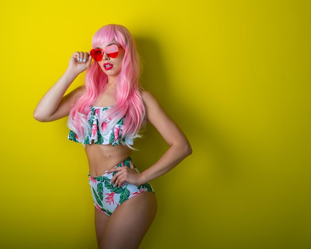 Schönes Mädchen mit rosa Perücke und farbigem Bikini posiert auf gelbem Hintergrund Frau mit künstlichen langen Haaren und rosa Brille in Form von Herzen