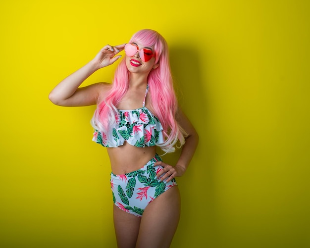 Schönes Mädchen mit rosa Perücke und farbigem Bikini posiert auf gelbem Hintergrund Frau mit künstlichen langen Haaren und rosa Brille in Form von Herzen