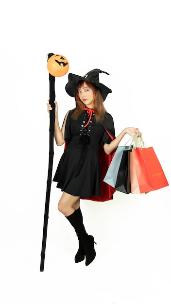 Schönes Mädchen mit schwarzen und orangefarbenen Haaren in schwarzem Kleid und Hexenhut hält Personal mit Einkaufstüten auf weißem Hintergrund für den Halloween-Tag