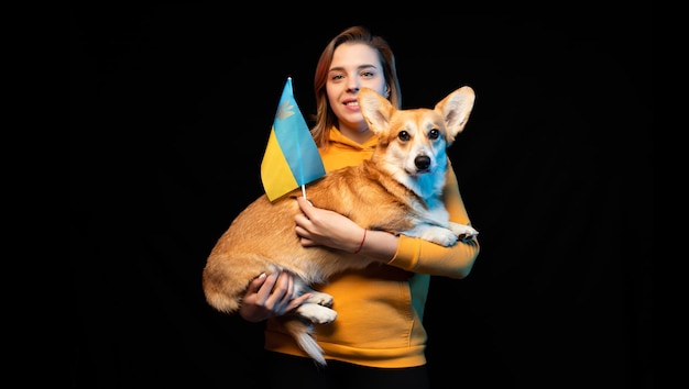 Schönes Mädchen mit Welsh Corgi Pembroke mit Ukraine-Flagge auf schwarzem Hintergrund Patriotischer ukrainischer Hund Friedlicher Protest gegen den Krieg
