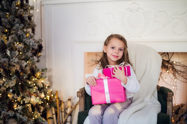 Schönes Mädchen nahe Weihnachtsbaum, das Geschenke auspackt, die auf einem Stuhl sitzen