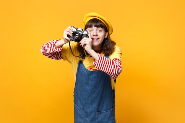 Schönes Mädchen Teenager in französischer Baskenmütze, Denim-Sommerkleid beim Fotografieren auf Retro-Vintage-Fotokamera isoliert auf gelbem Wandhintergrund. Menschen aufrichtige Emotionen Lifestyle-Konzept. Mock-up-Kopienbereich.